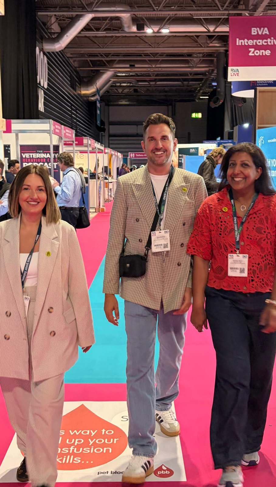 Three people smiling and walking at a trade show with pink and blue carpeted aisles.