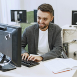 Man smiling while typing at a desktop computer in an office with papers on the desk