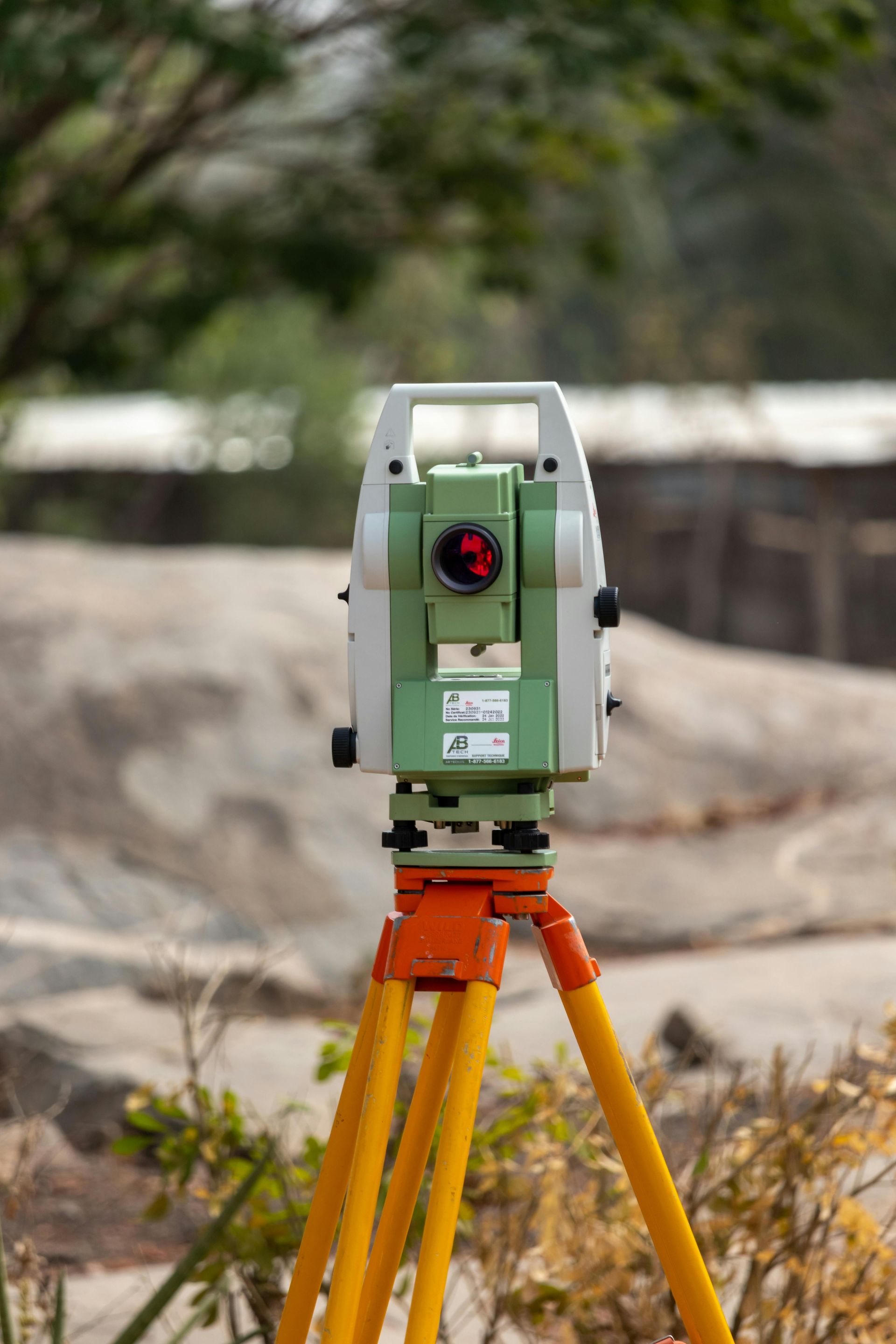 Total station surveying instrument mounted on a tripod, outdoors. Orange, yellow, and green colors are prominent.