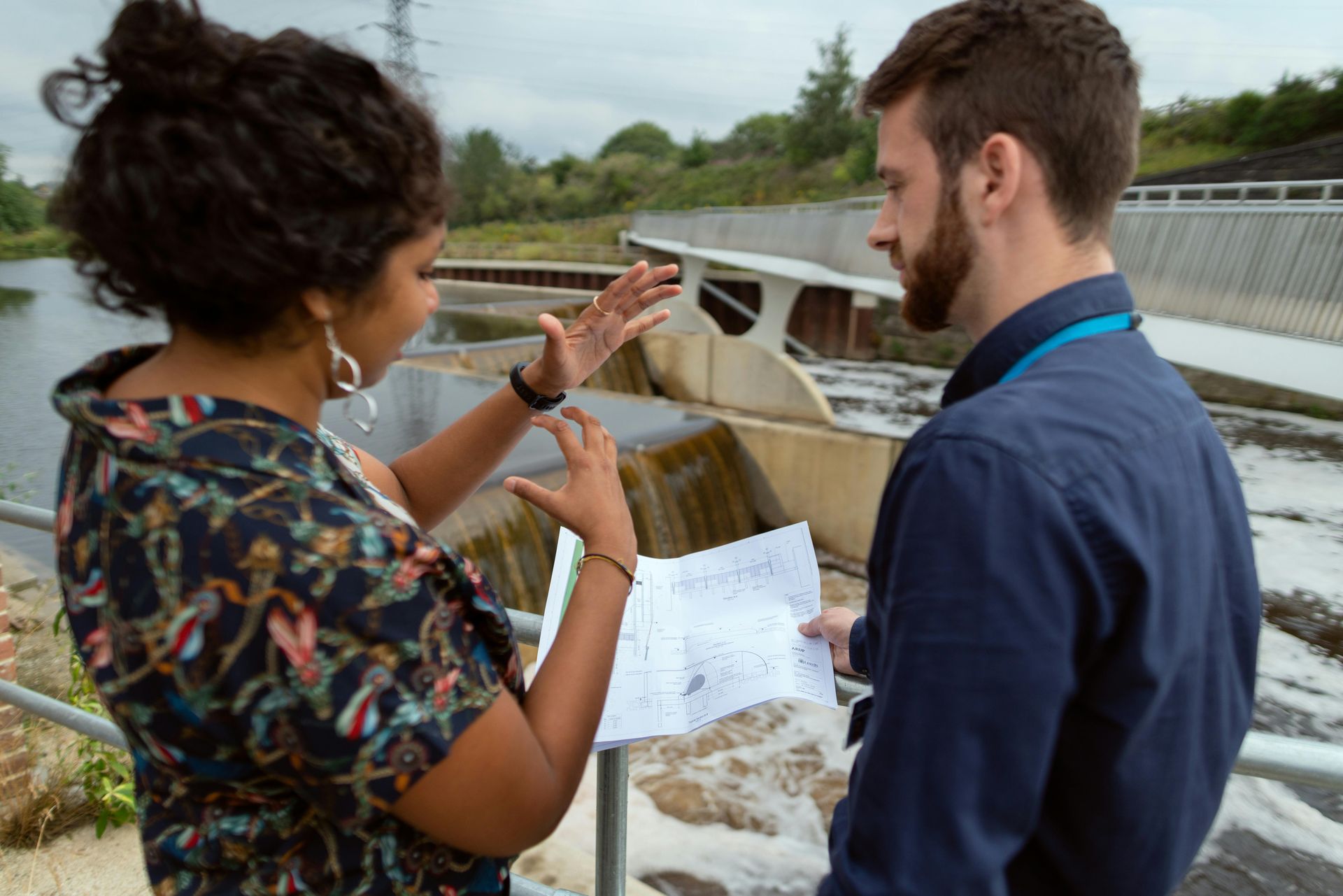Two people looking at papers by a dam, discussing. One points with hands.