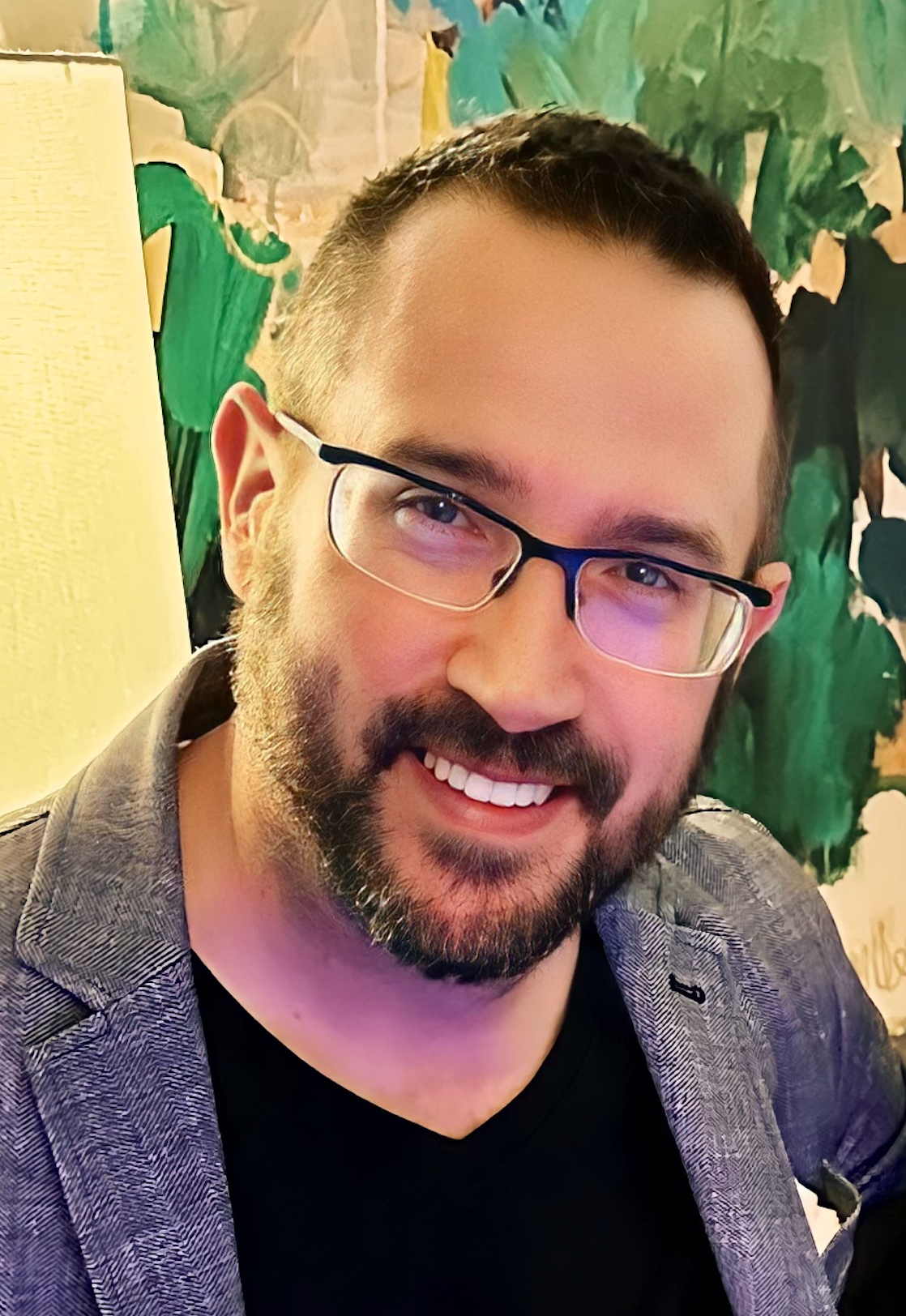 A man with a beard and glasses is smiling in front of a painting.