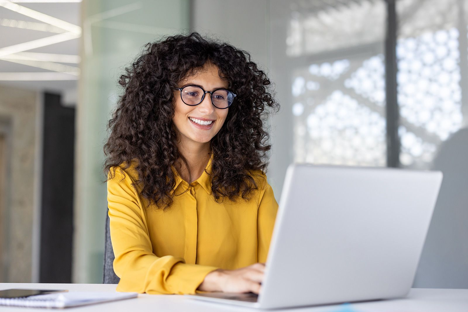 Smiling person in a yellow shirt typing on a laptop in a bright office