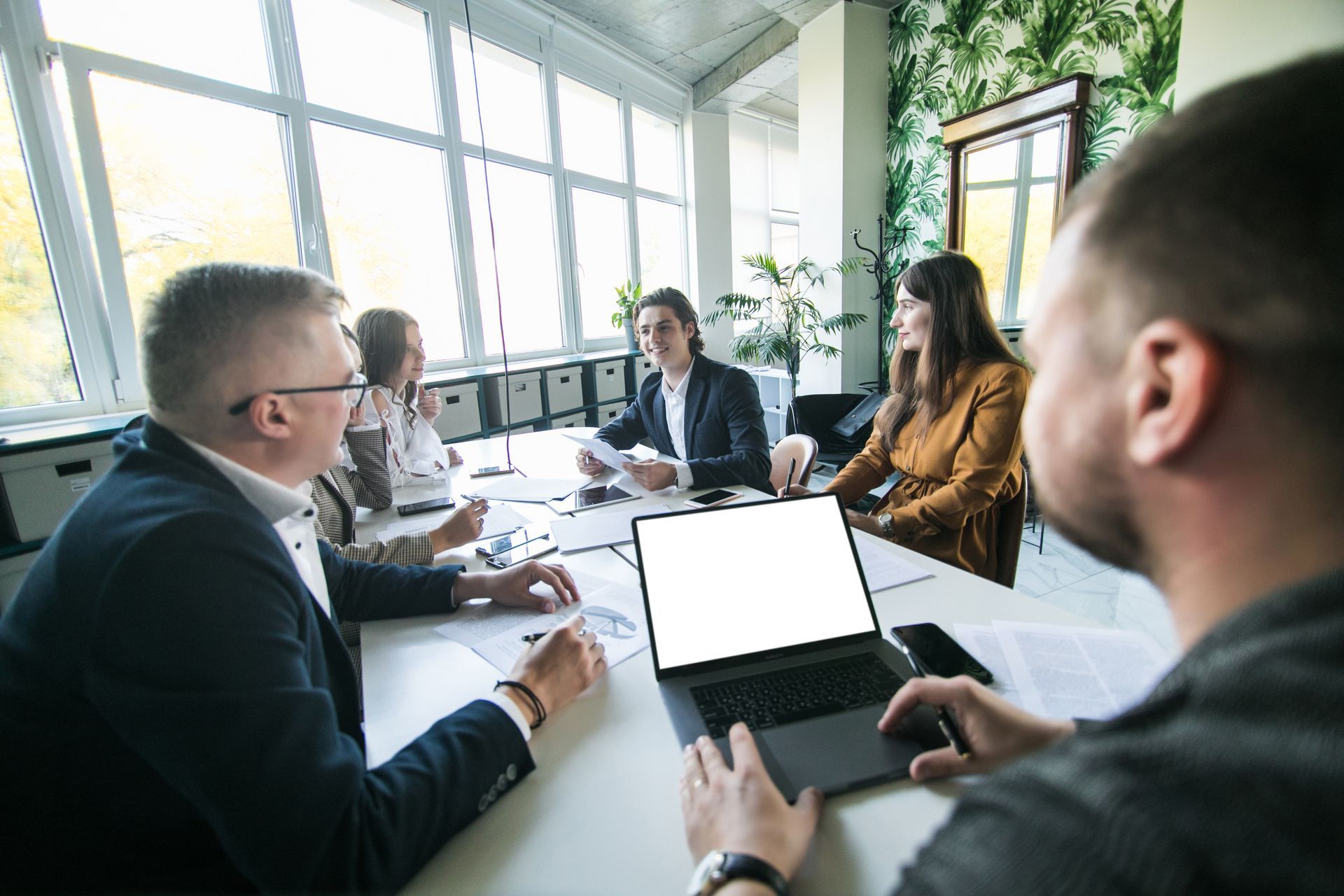 Business meeting around a conference table in a bright office, with laptops and papers visible.