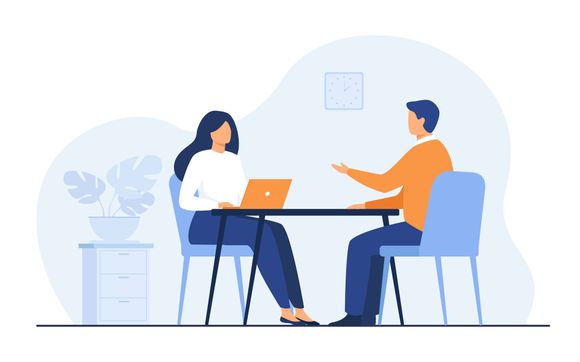 Two people sit at a table in a bright office, talking with a laptop and a plant nearby.