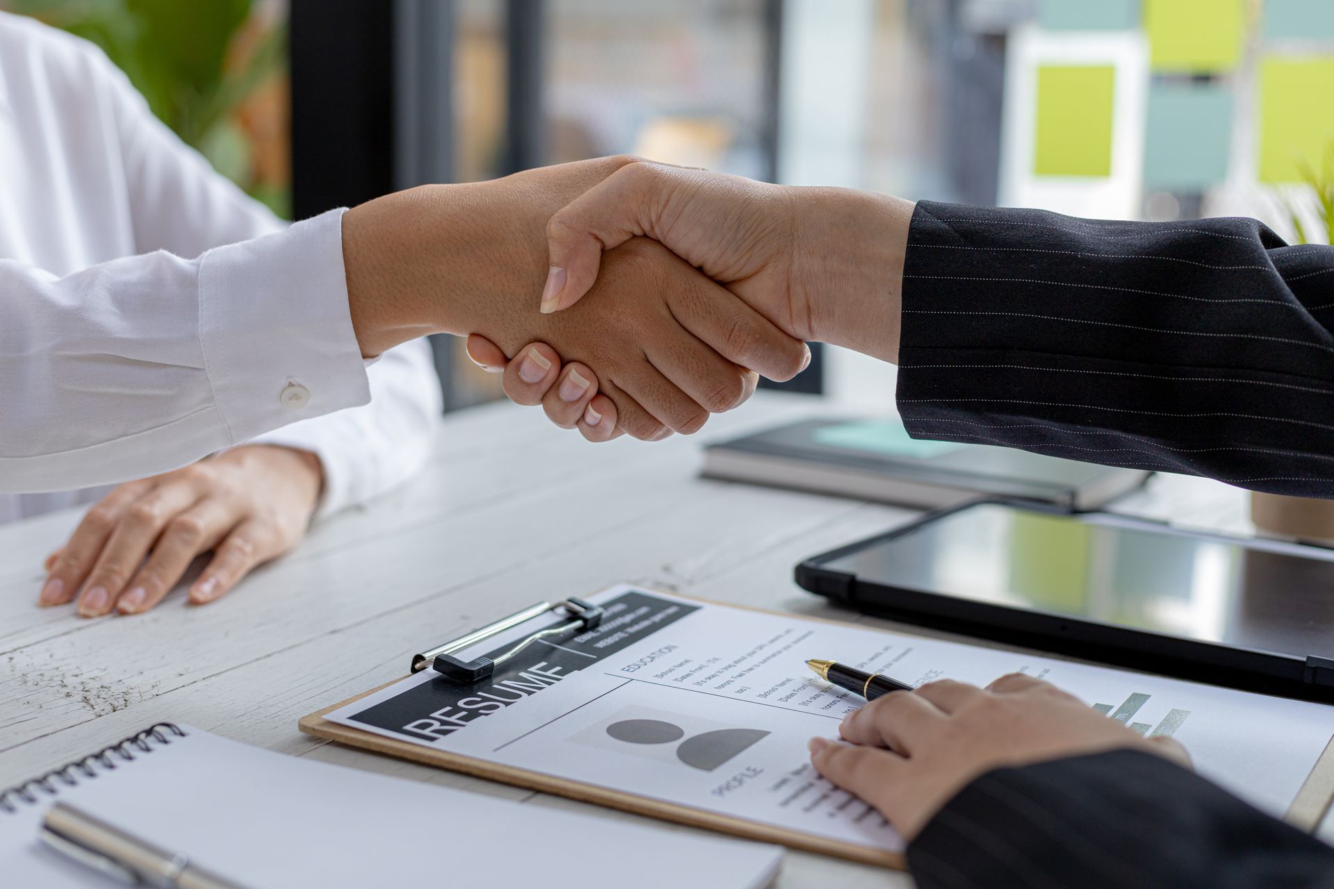 Business handshake across a desk with resume clipboard, tablet, and notebook.