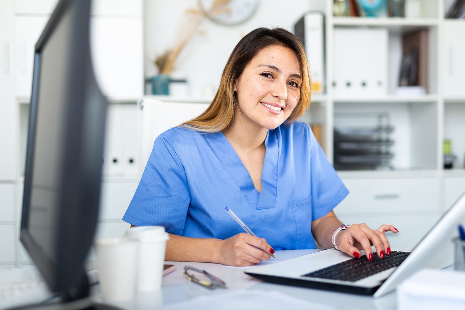 Smiling woman in blue scrubs writing at a desk with a laptop in an office