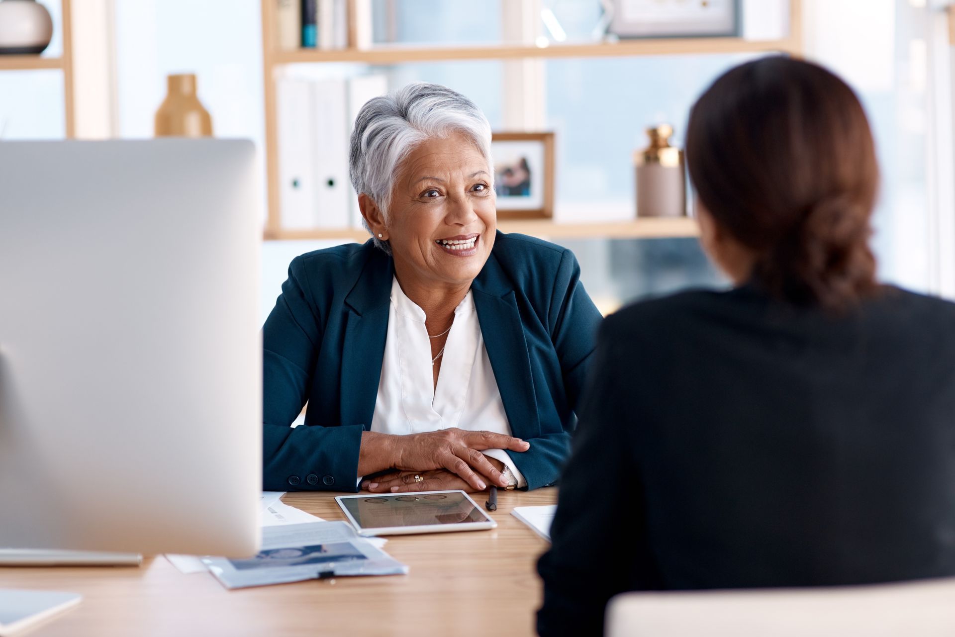 Two people in a bright office meet across a desk, one smiling and speaking to the other.