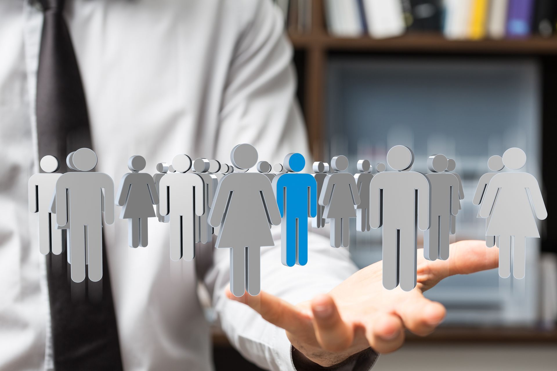 Highlighted blue human figure among gray people icons hovering above an open hand. Highlighted blue human figure among gray people icons hovering above an open hand.