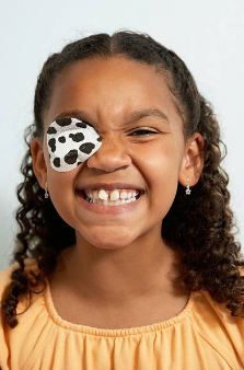 Girl with curly hair wearing a cow-print eye patch, smiling and squinting against a light-colored background.