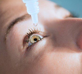 A person's eye receiving eye drops from a white bottle.