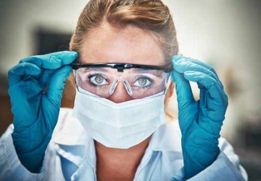 Scientist in a lab coat, mask, and gloves adjusting safety goggles; intense focus.