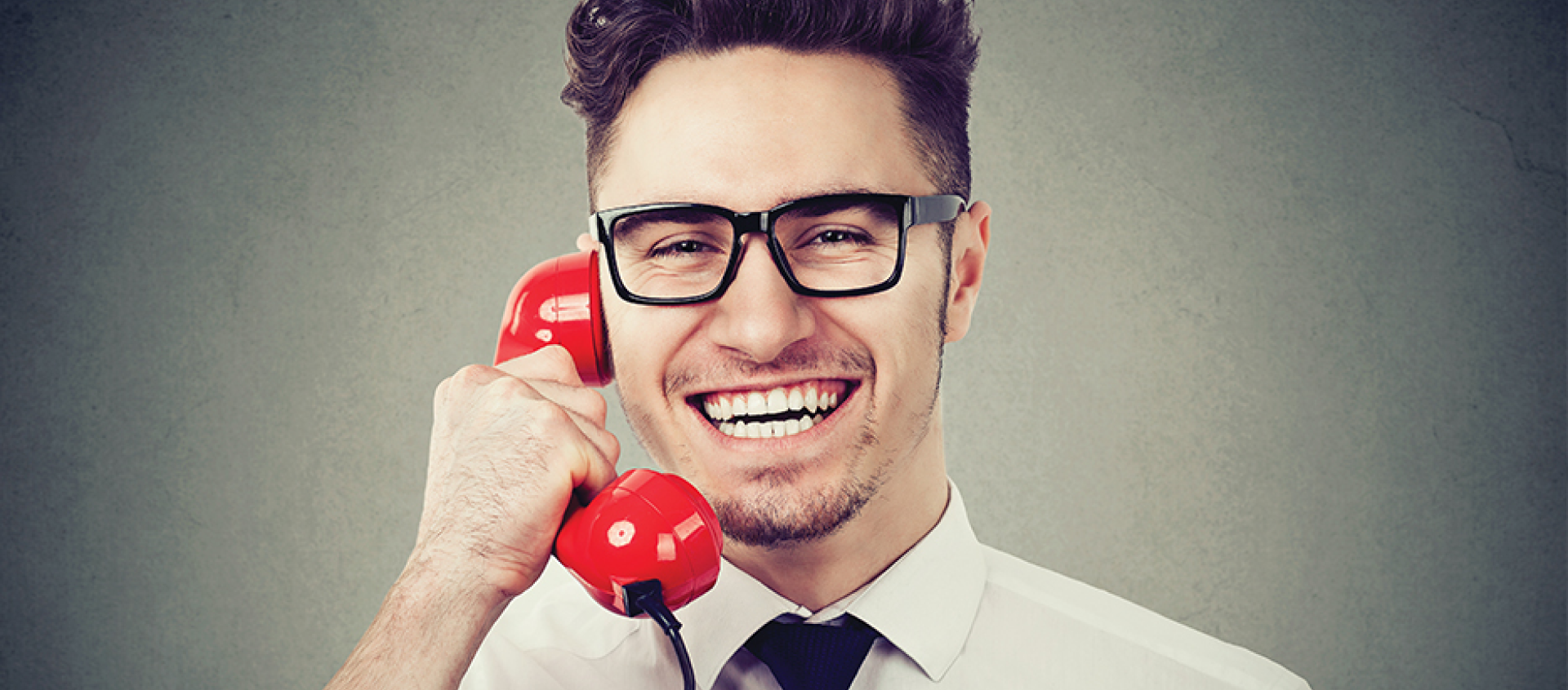 A man wearing glasses is smiling while talking on a red telephone.