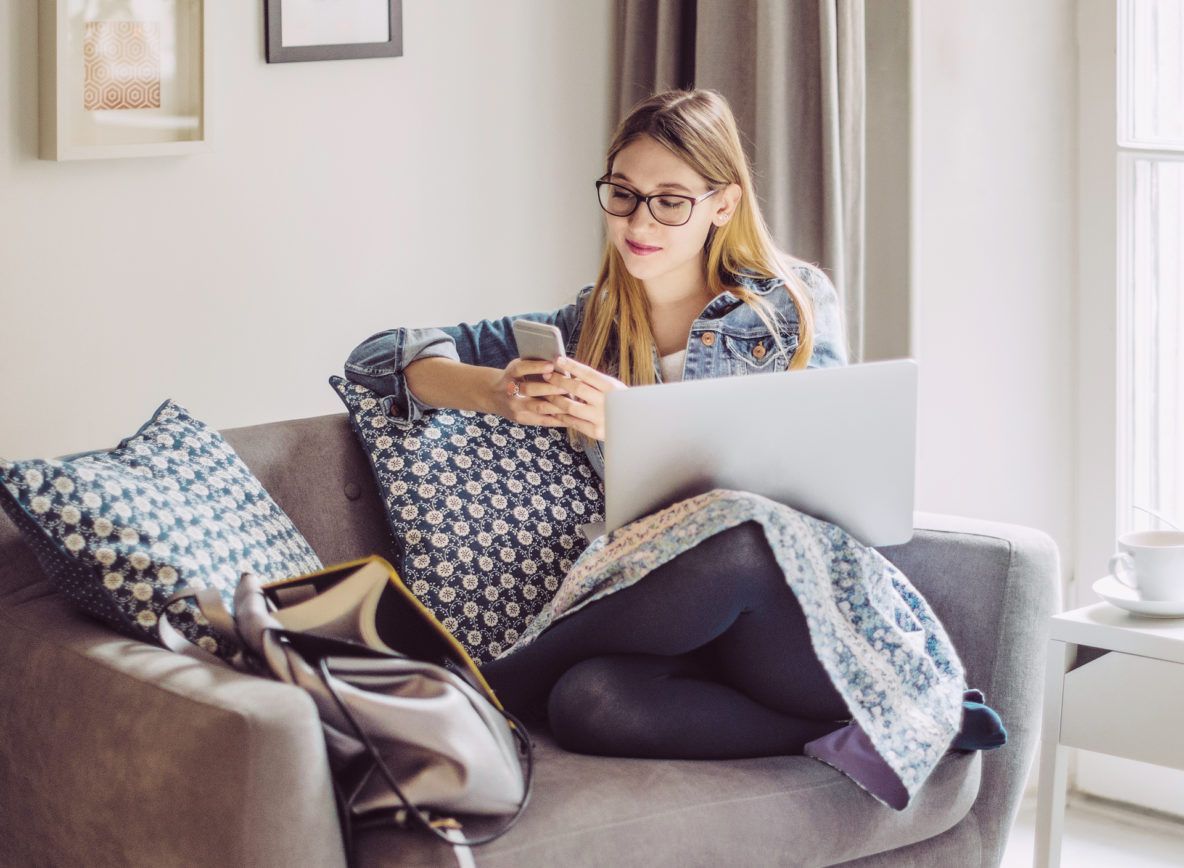 A woman is sitting on a couch using a laptop and a cell phone.