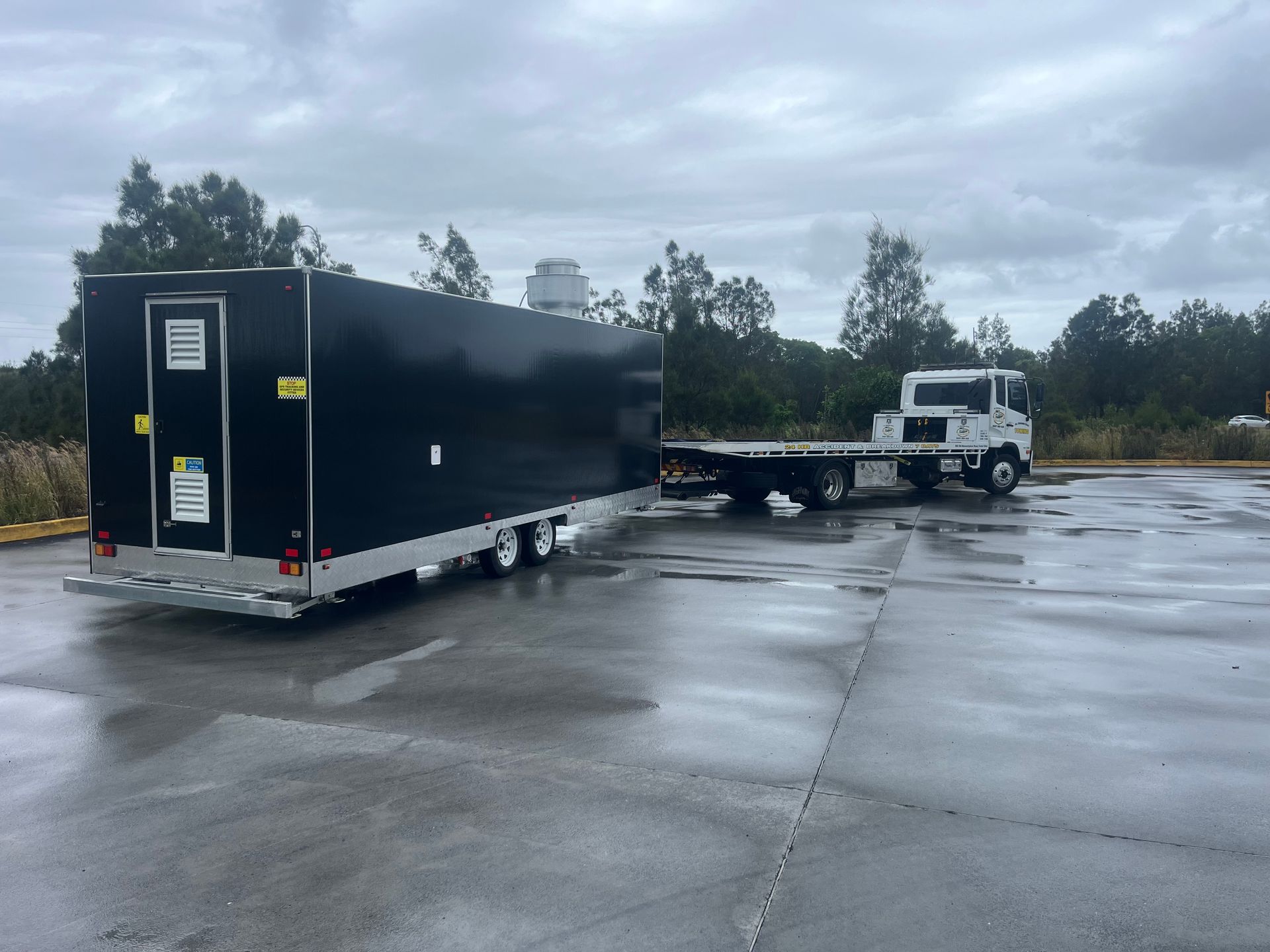 White truck towing a black enclosed trailer across a wet parking lot under cloudy skies — All Sunshine Coast Towing In Coolum, QLD