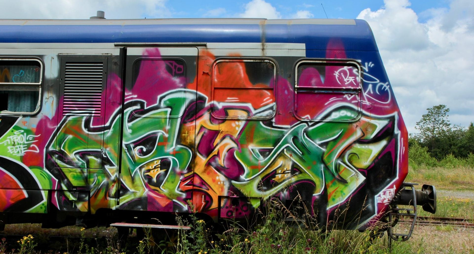 Ein verwitterter Eisenbahnwaggon auf einem grasbewachsenen Feld, dicht bedeckt mit leuchtend grünen, orangen und violetten Graffiti-Schriftzügen.