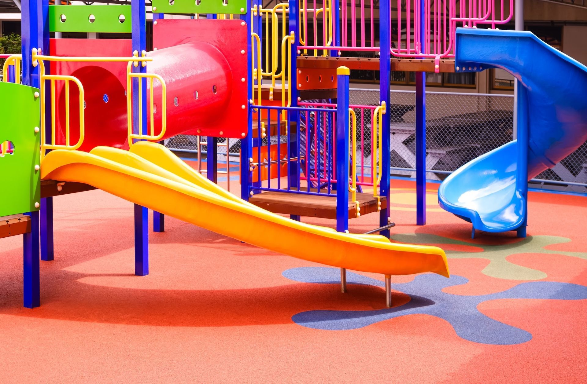 Ein farbenfroher Spielplatz mit einer doppelten gelben Rutsche, einem roten Tunnel und einer blauen Spiralrutsche auf rotem Gummiboden.