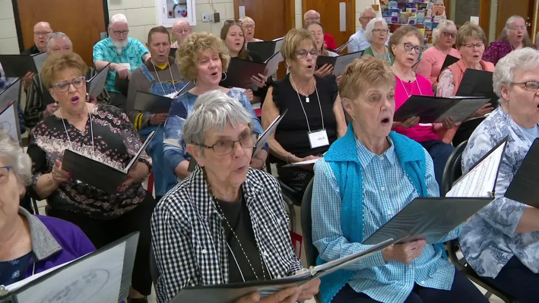 Choir members seated indoors, singing from sheet music in a bright room
