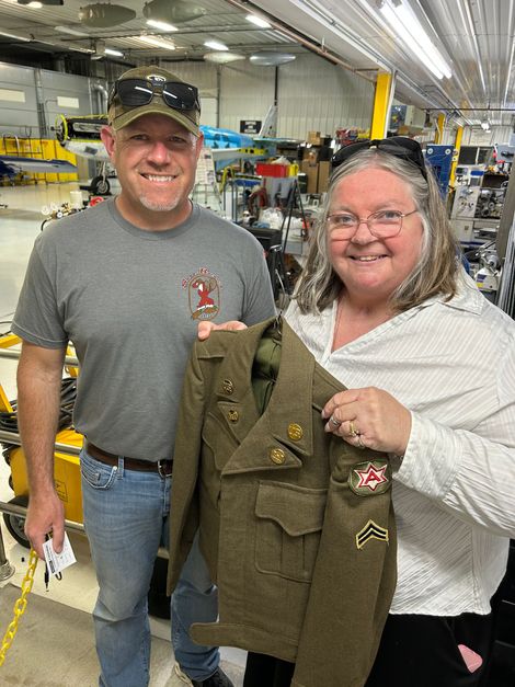 Man and woman holding an olive drab military jacket in a factory. They are smiling.