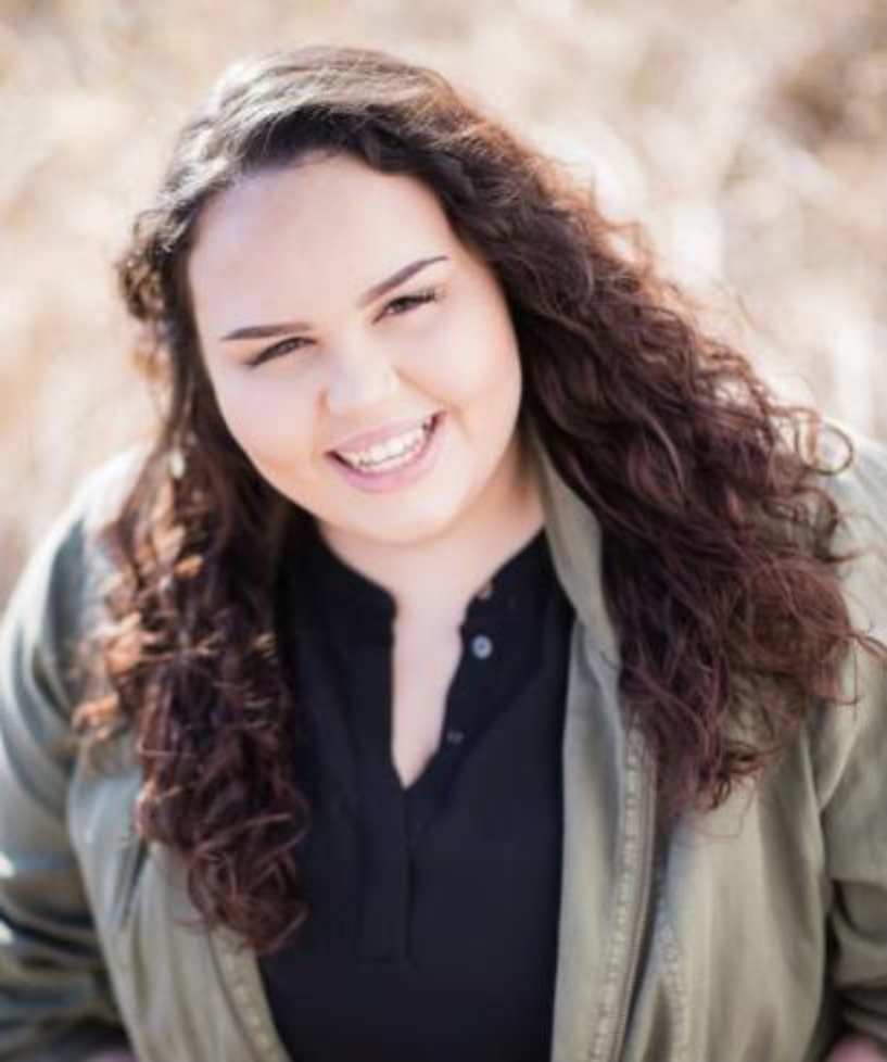 A smiling person with long, curly dark hair wearing a black shirt and a light olive green jacket outdoors.