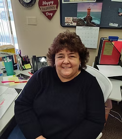 Woman in black sweater at a desk, smiling. Office setting with calendar and Valentine's Day decor.