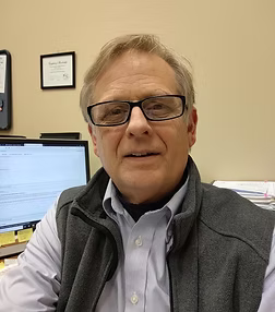 Man in glasses and vest smiles, sitting at a desk with a computer.