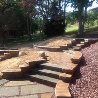 Curved stone garden path lined with flowers and ferns leading to a stone bench in a sunny backyard