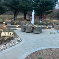 Curved stone patio with outdoor seating, lanterns, and lush landscaped garden under a cloudy sky