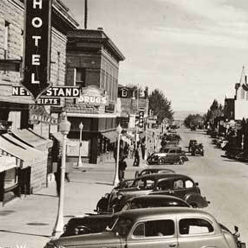 A black and white photo of a city street with cars parked on the side of the road. Historic Downtown Rawlins.