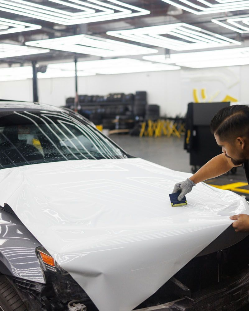Two people applying white protective film to a car hood inside a well-lit shop.