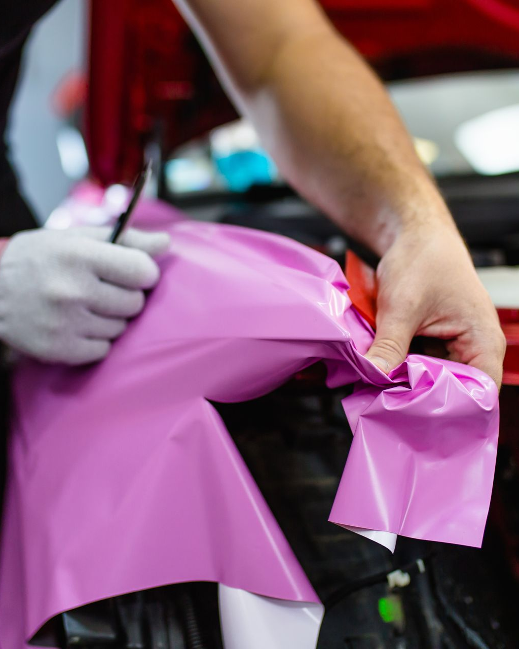 A person in gloves wrapping a pink vinyl sheet on a car's bumper using a blade.