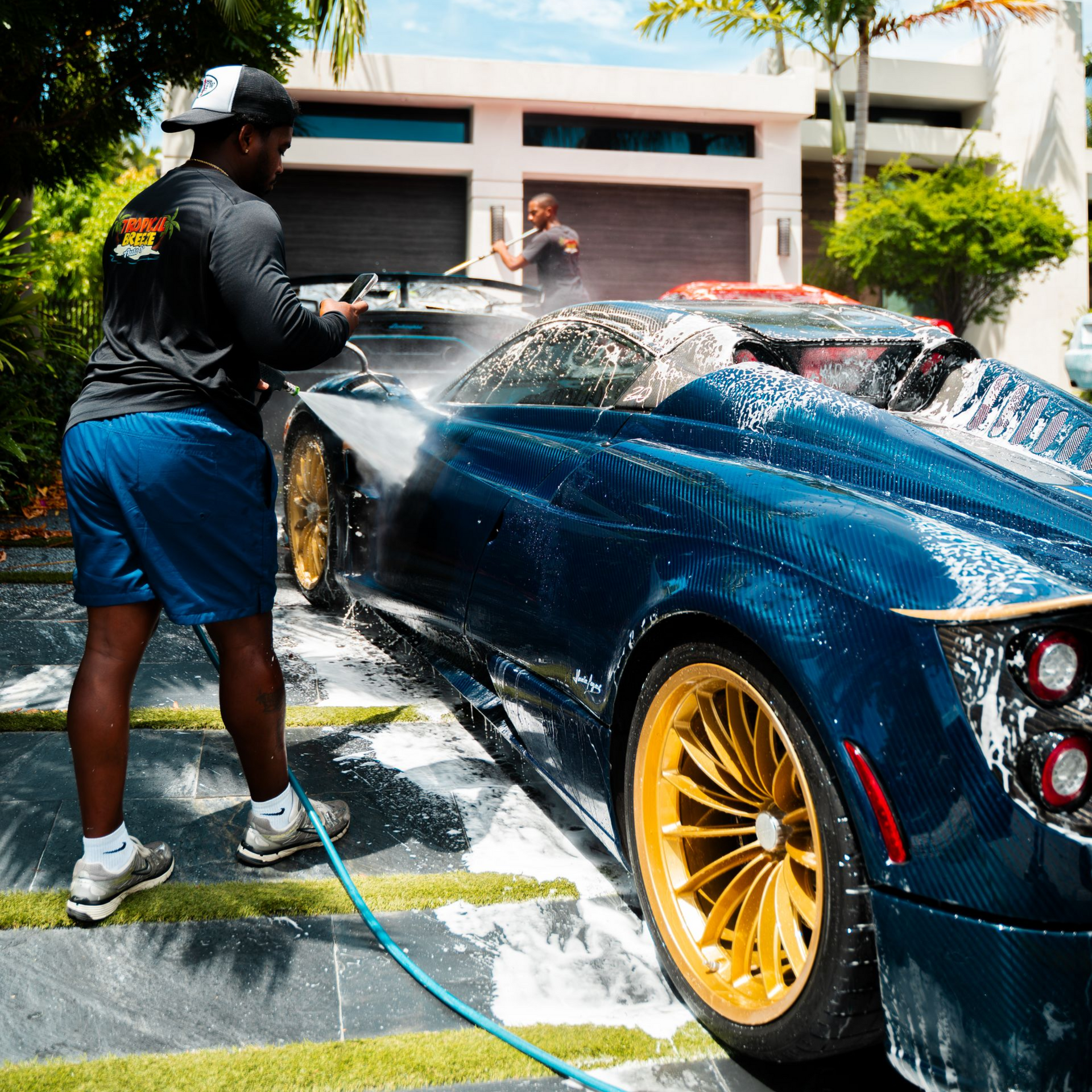 Person washing a dark blue Pagani Huayra with gold wheels in front of a modern house.