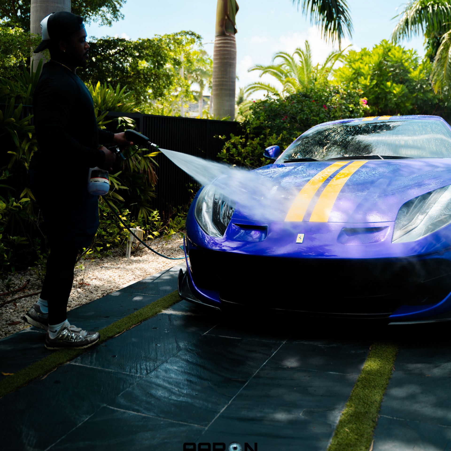 Person washing a blue Ferrari with yellow stripes outdoors in a sunny setting.