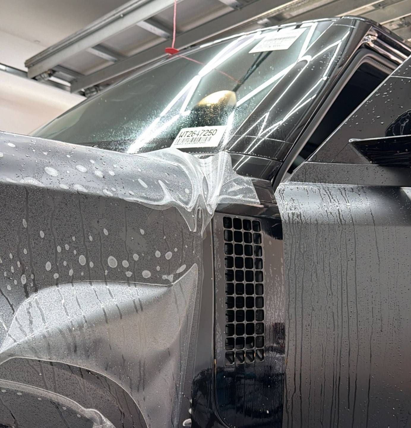 Black car in a garage, covered in soap suds with water droplets on the hood and windshield.