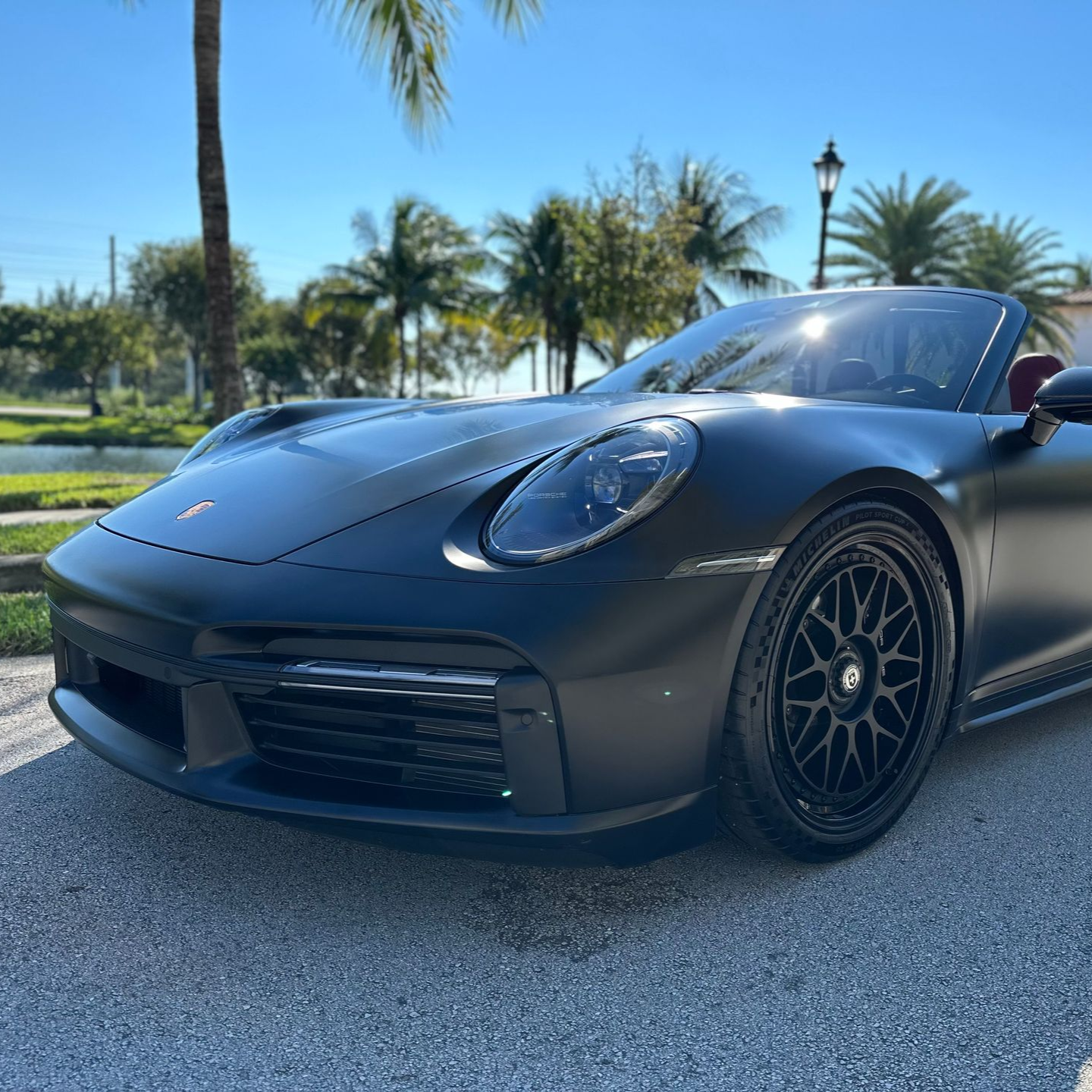 Matte black Porsche convertible parked on a sunny street, palm trees in the background.