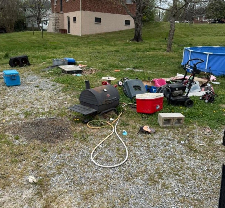 Scattered yard tools and equipment on gravel near a house and lawn, including a grill, bins, and a blue kiddie pool