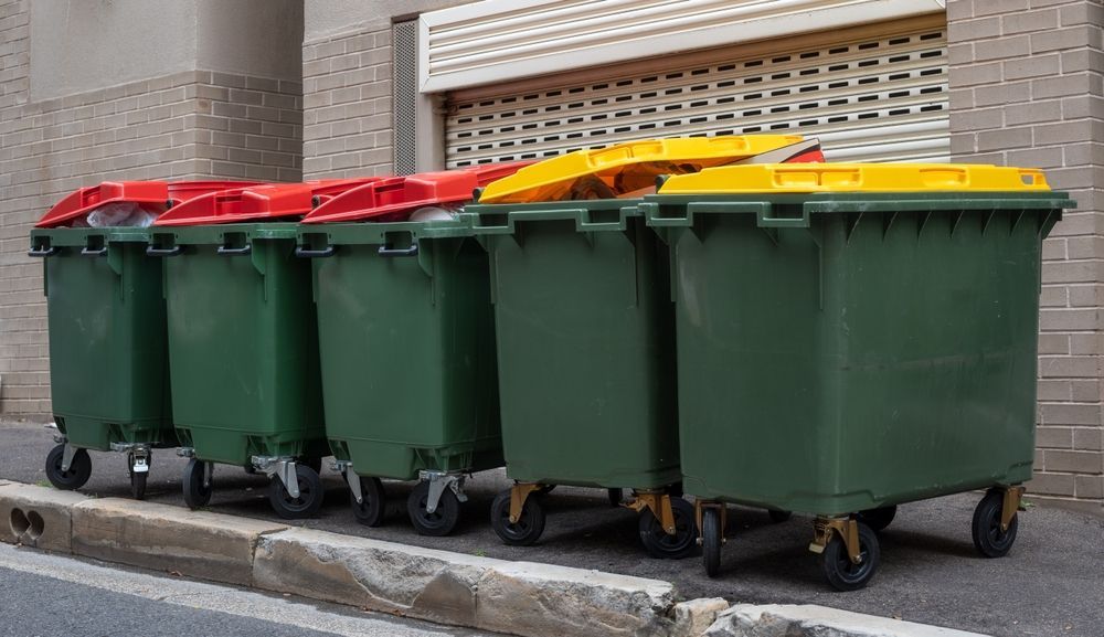 Row of green wheeled trash bins with red and yellow lids lined up on a street curb