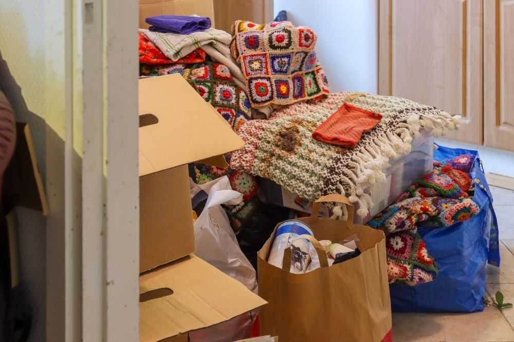 Cluttered room with stacked boxes, bags, folded blankets, and cushions near a doorway.