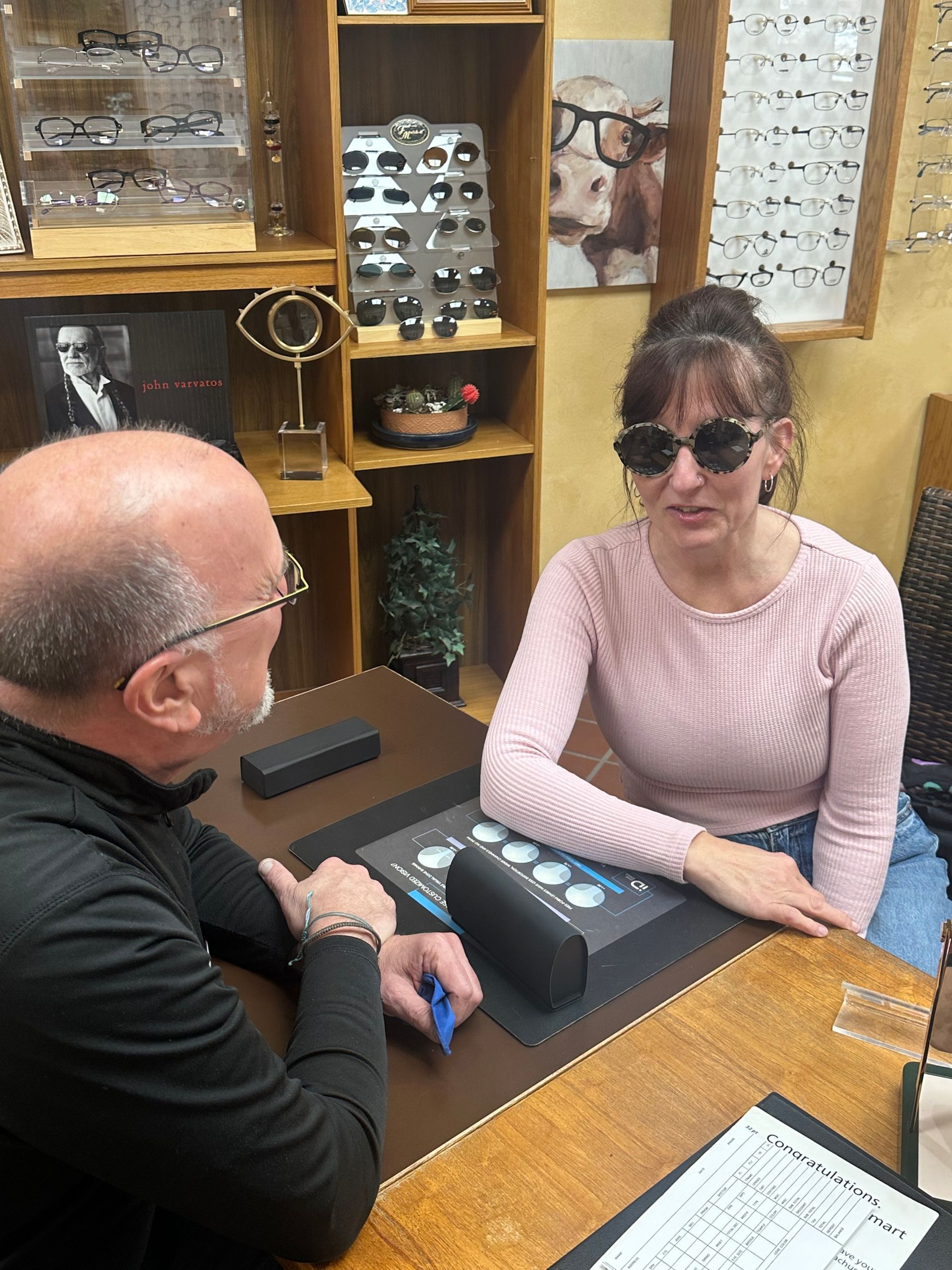 A woman wearing sunglasses sits opposite a person at a table, discussing eyewear options in a store.
