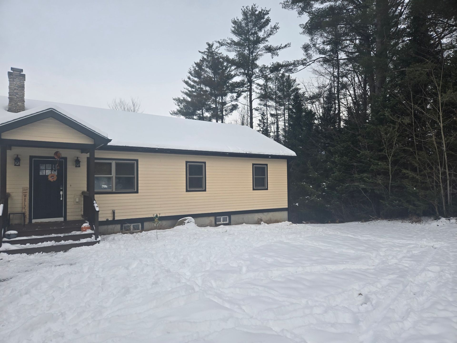 Beige house with snow-covered roof and yard; trees in the background. Winter scene.