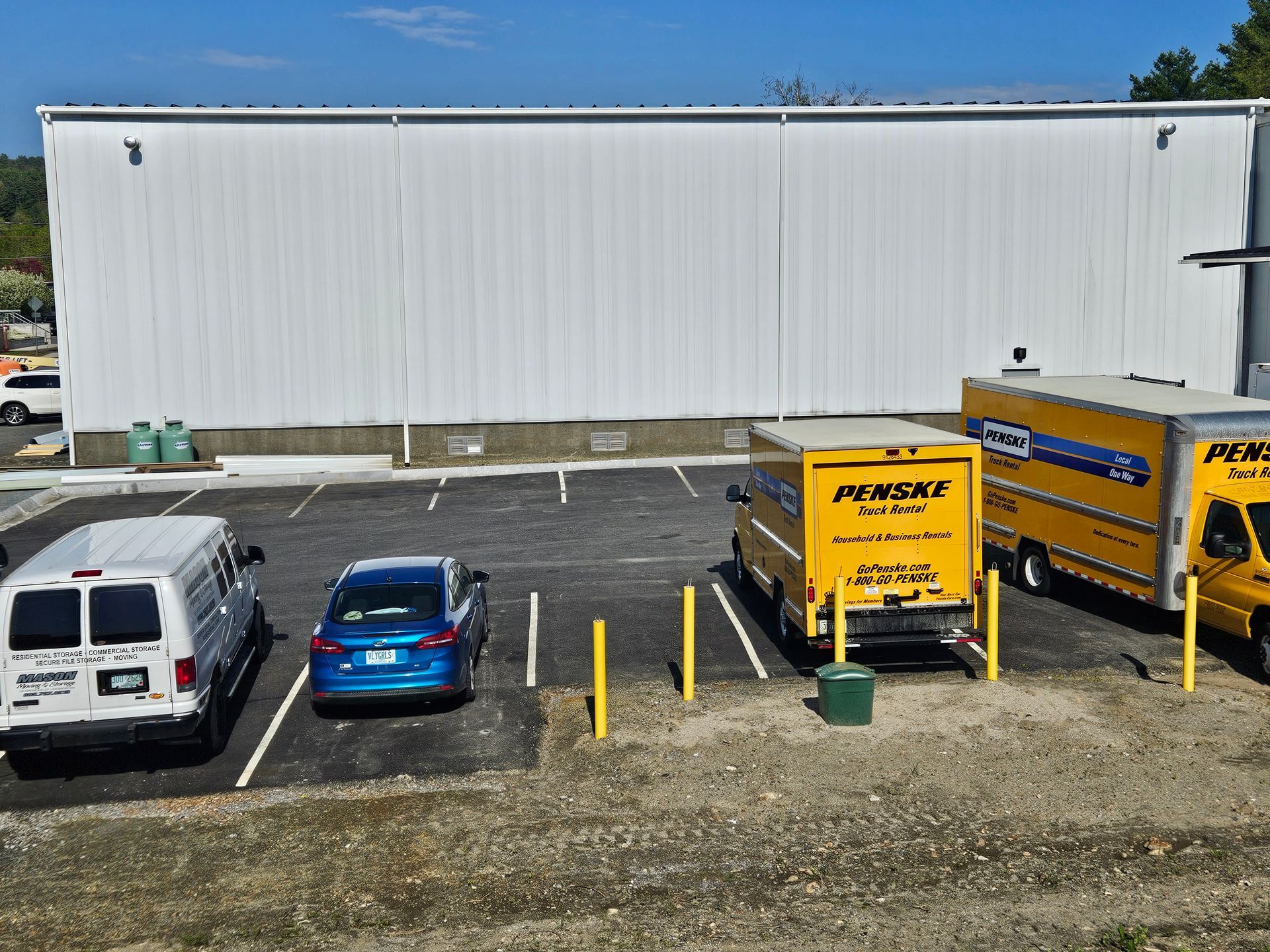 Penske trucks and vans parked in front of a large white building, blue car also parked. Sunny day.