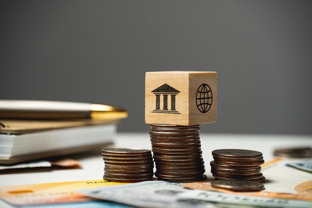 Stacks of coins with wooden blocks showing bank and globe icons on a desk, symbolizing finance and global trade