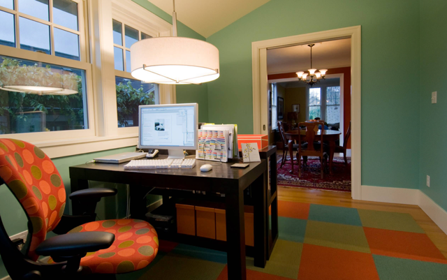 Home office with desk, computer, lamp, orange chair, and colorful rug by a window and open doorway