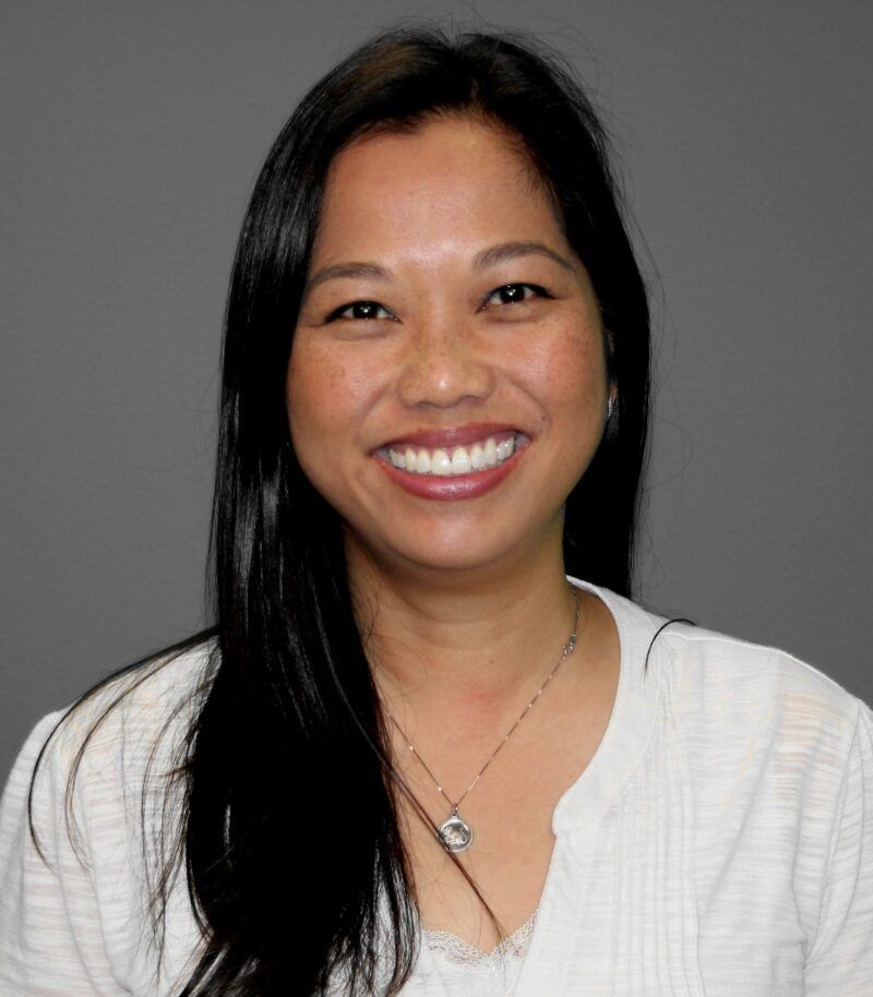 Woman smiling with long dark hair and light colored top gray background