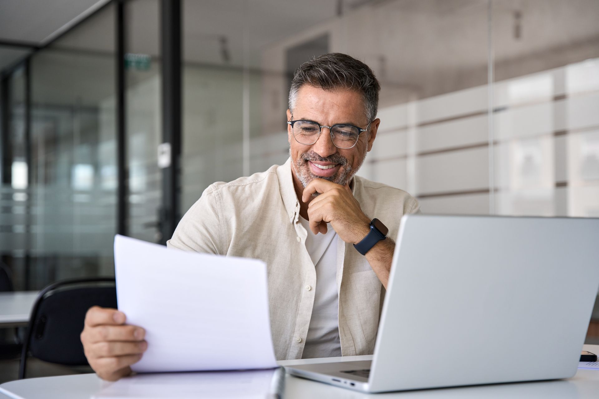 Un homme portant des lunettes regarde un document, son ordinateur portable est ouvert sur son bureau.