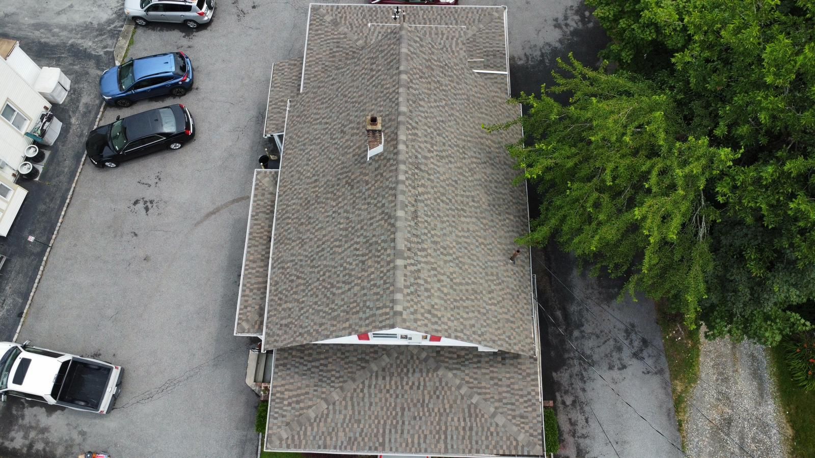 Aerial view of a building with a gray shingle roof, chimney, and surrounding parking area with vehicles and green trees.