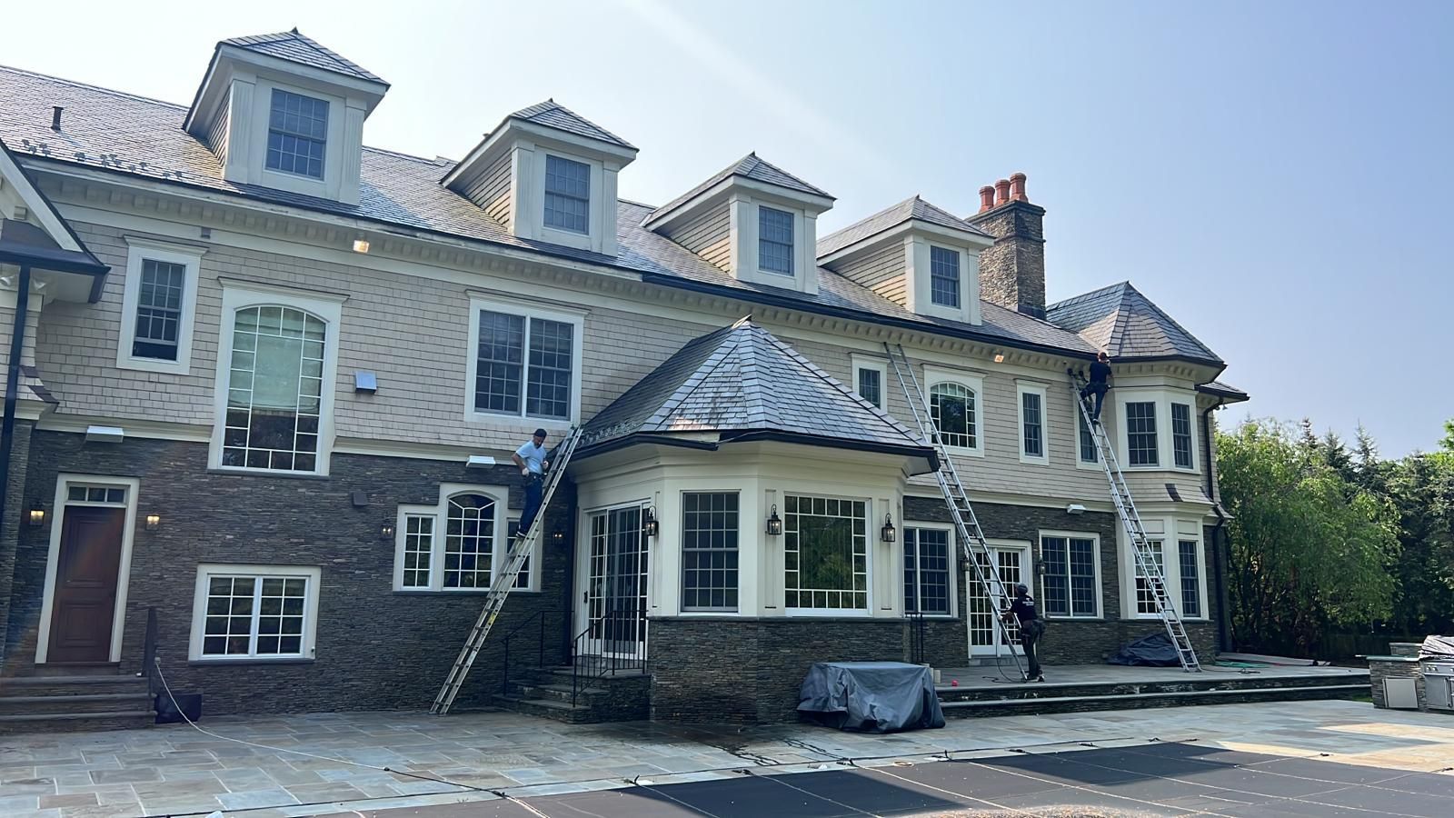 Workers on ladders painting the exterior of a large, two-story house with dark brick and light-colored siding.