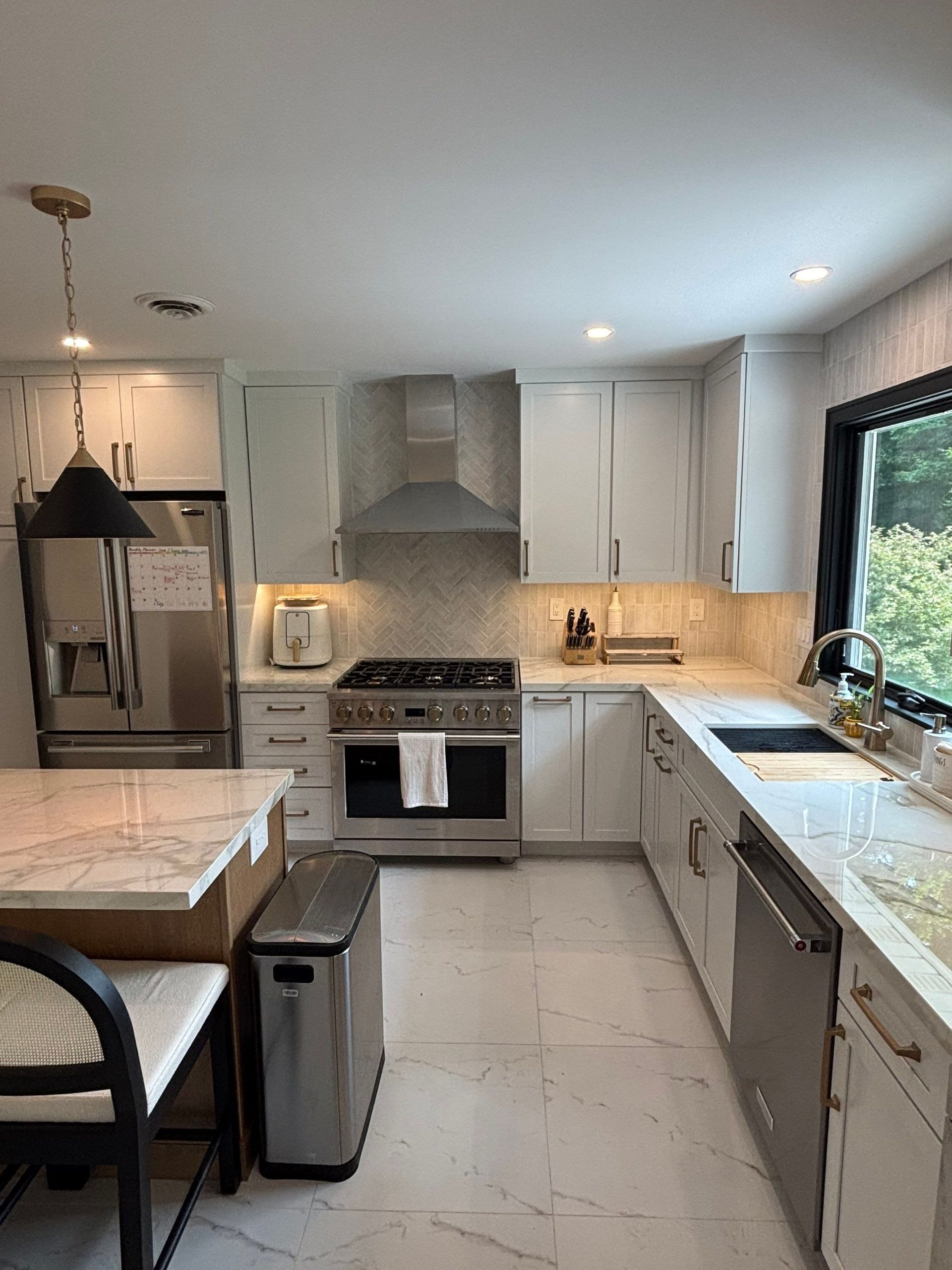 Modern white kitchen with stainless steel appliances, marble countertops, and a large window.
