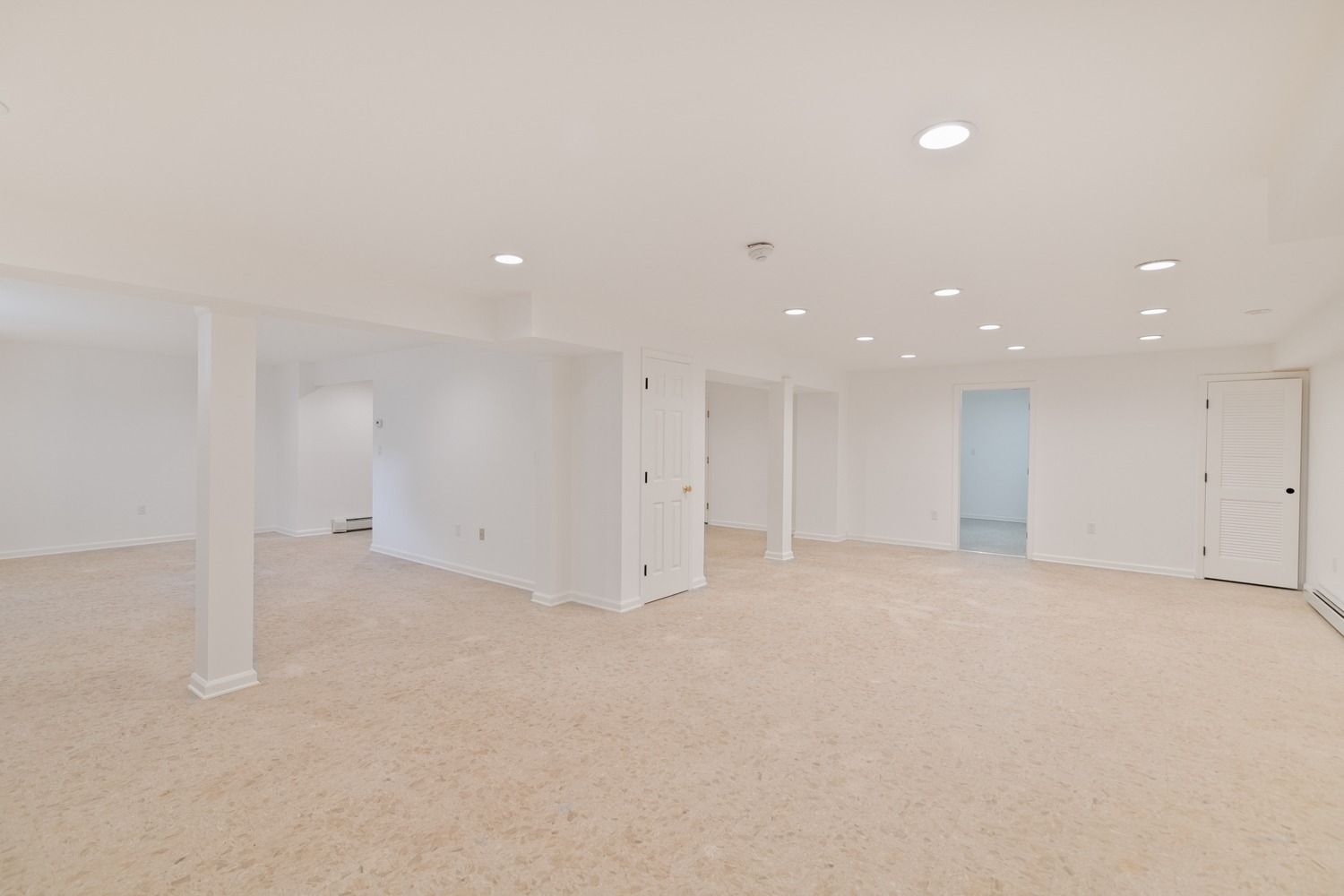 Empty, bright white basement with beige carpet and several support columns and doorways.