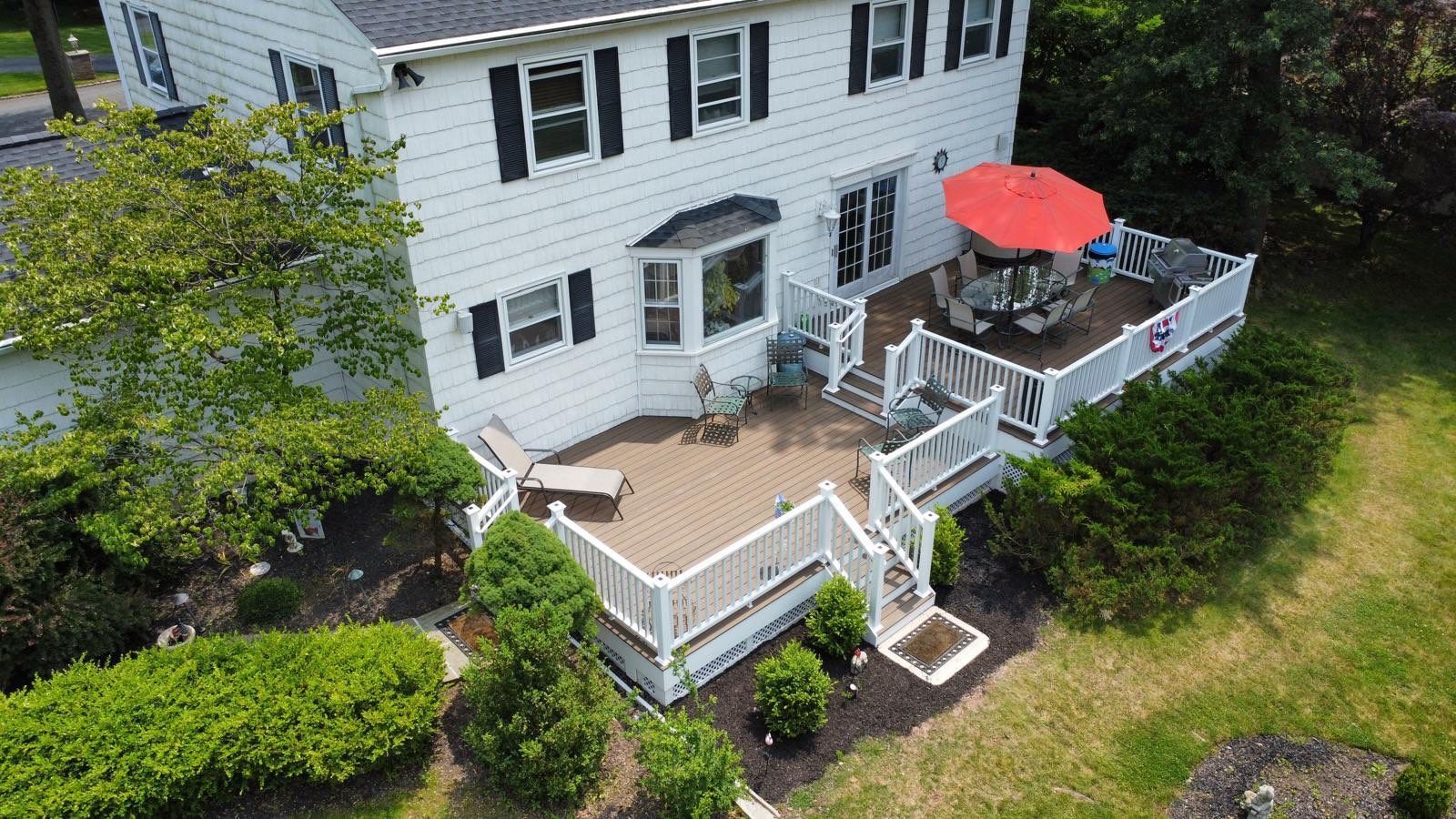 White house with a brown deck, red umbrella, and greenery.