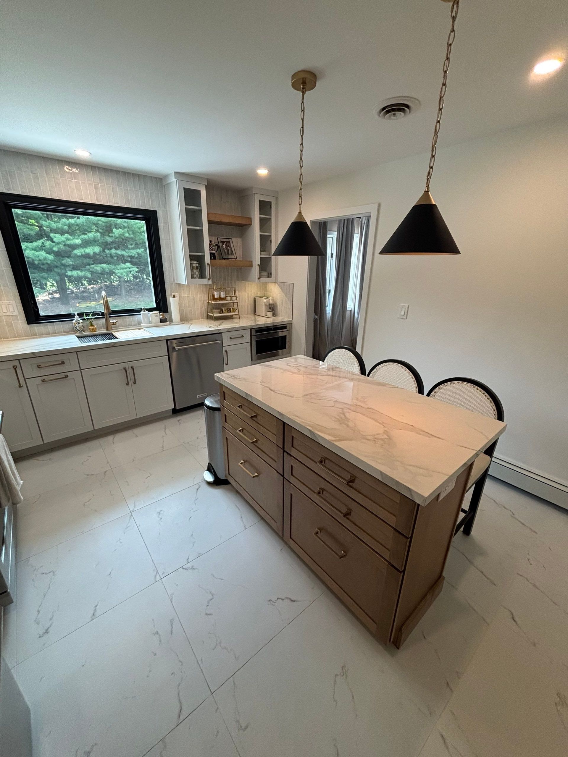 Kitchen with island, light cabinets, black pendant lights, large window, and light-colored countertops and flooring.
